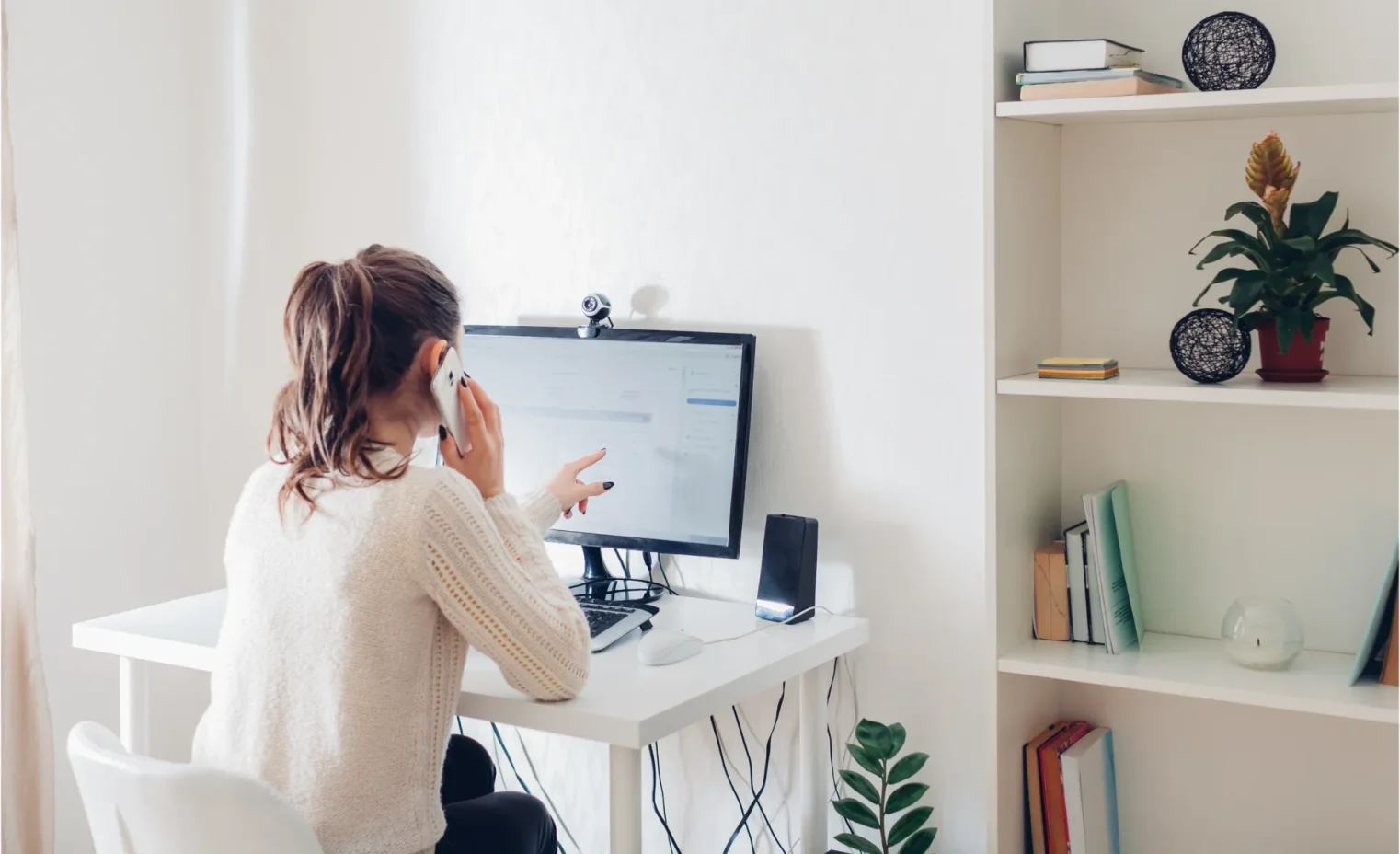 a woman working from a desktop computer talking on a smartphone in a home office.webp