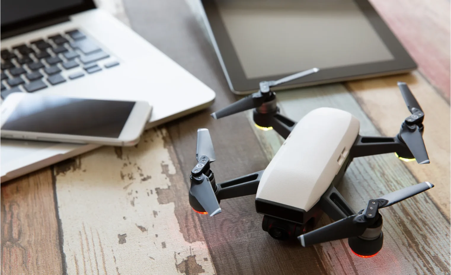 a drone switched on beside a macbook laptop and a tablet on a wooden office desk.webp